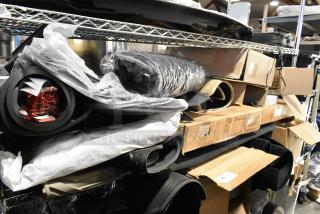 Lot of various items including car floor mats, rolled cloths, and boxed goods on metal shelves in a warehouse setting.