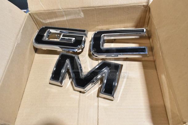 Alt text: "GM logos in a cardboard box from a lot including wheel locks and a stick shift in black poly bin."