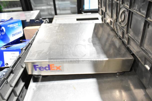 A FedEx metal scale on a shelf, surrounded by Samsung boxes and a black poly bin. Scale shows signs of wear.