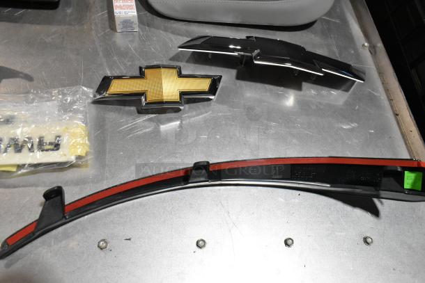 Lot of various automotive emblems and parts, including Chevrolet logo, on metal surface. Items in used condition.