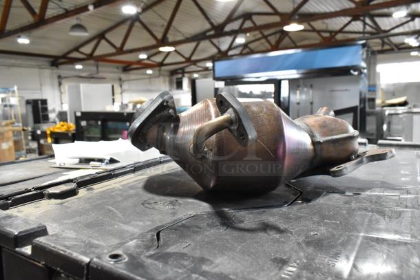 Metal automotive part, possibly a catalytic converter, with mounting flanges, in used condition, on a workshop table.