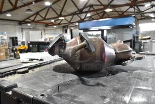 Metal automotive part, possibly a catalytic converter, with mounting flanges, in used condition, on a workshop table.