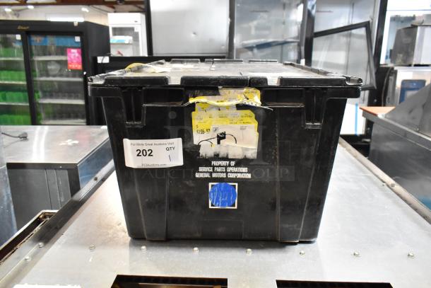 Black poly bin containing a lot of various items, including GM front driver side fender ornament 20823543. Visible labels note auction info and General Motors Corporation ownership.