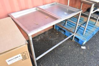 Stainless steel table with dual surfaces, some wear visible, sturdy metal legs, suitable for commercial use.