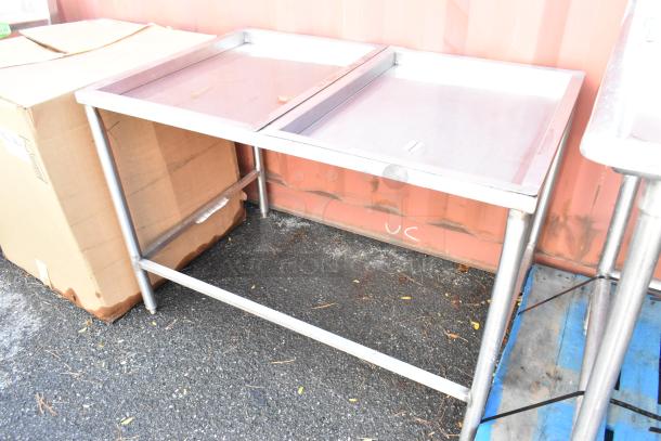 Stainless steel table with dual trays and tubular legs, shown outdoors, in used condition. Ideal for catering or industrial use.