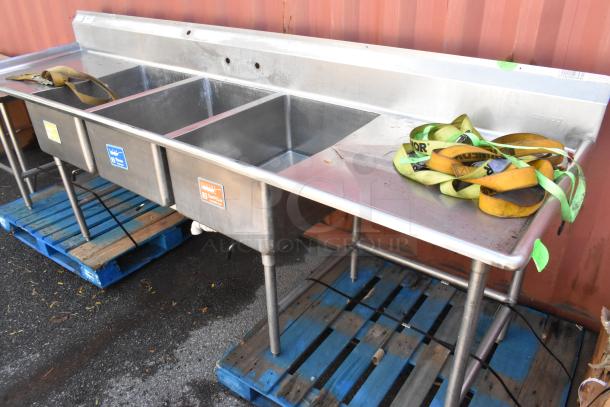 Stainless steel 3 bay sink with dual drain boards, used condition. Labels indicate wash, rinse, sanitize.