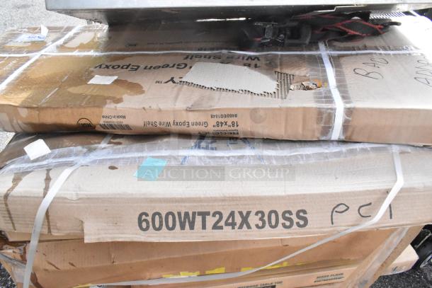 Stack of packaged stainless steel work tables and mop sink, some with visible wear on packaging, marked as epoxy wire shelves and table tops, labeled 600WT24X30SS.