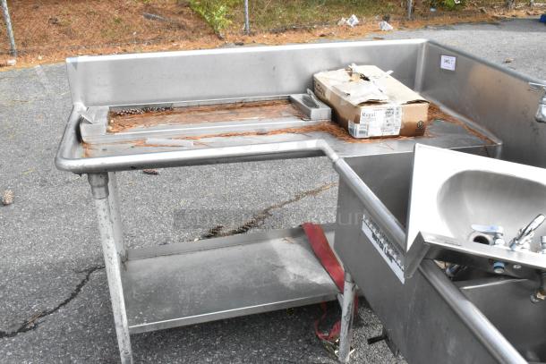 Stainless steel commercial L-shaped 3-bay sink with dual drain boards and under shelf, showing signs of outdoor storage.