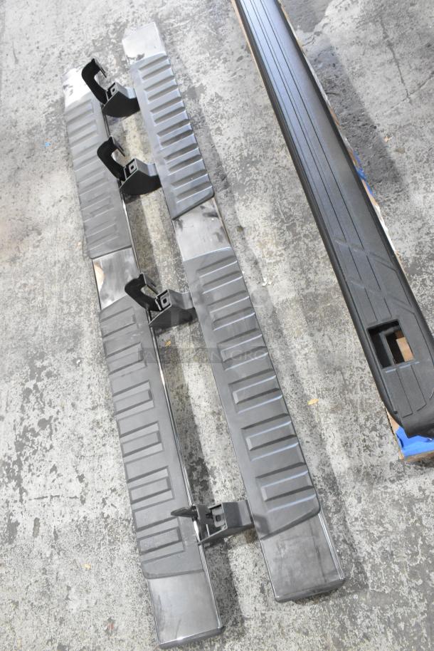 Four car parts, including step bars and brackets, laying on concrete. Condition appears used. No visible labels.