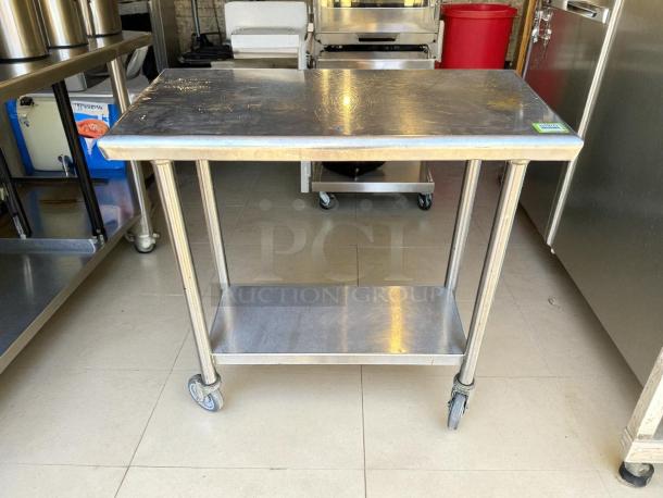 Stainless steel worktop table on casters with rounded edges, excellent condition, lower shelf, and label visible on top.