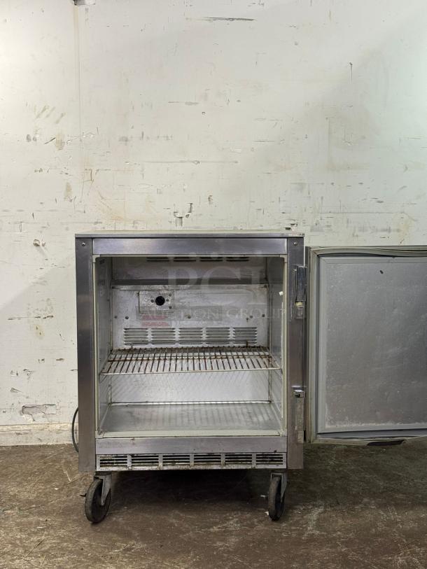 Under counter 28" refrigerator on wheels, stainless steel, open door showing wire shelf, signs of wear, ideal for compact spaces.