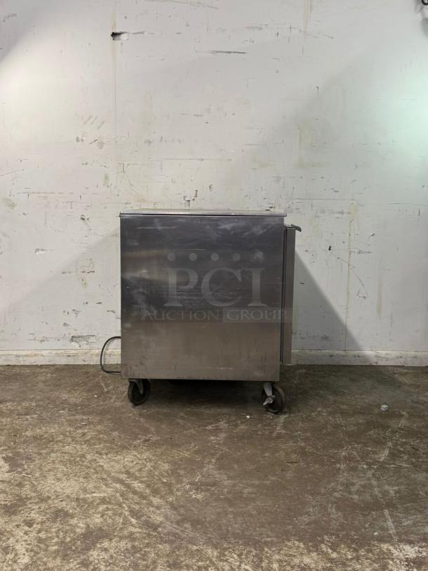 Stainless steel under-counter 28" refrigerator on wheels, showing signs of wear, suitable for compact spaces.
