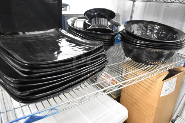 Black plates, bowls, and poly bin on a metal shelf. Items appear in good condition. Shelving unit not included.