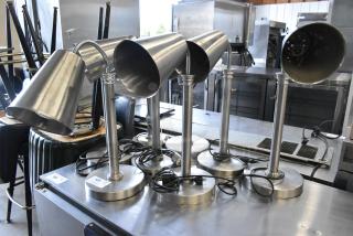 Eight metal heat lamps, 115 volts, 1 phase, on a counter. Includes cords. Auction lot tags visible at base.