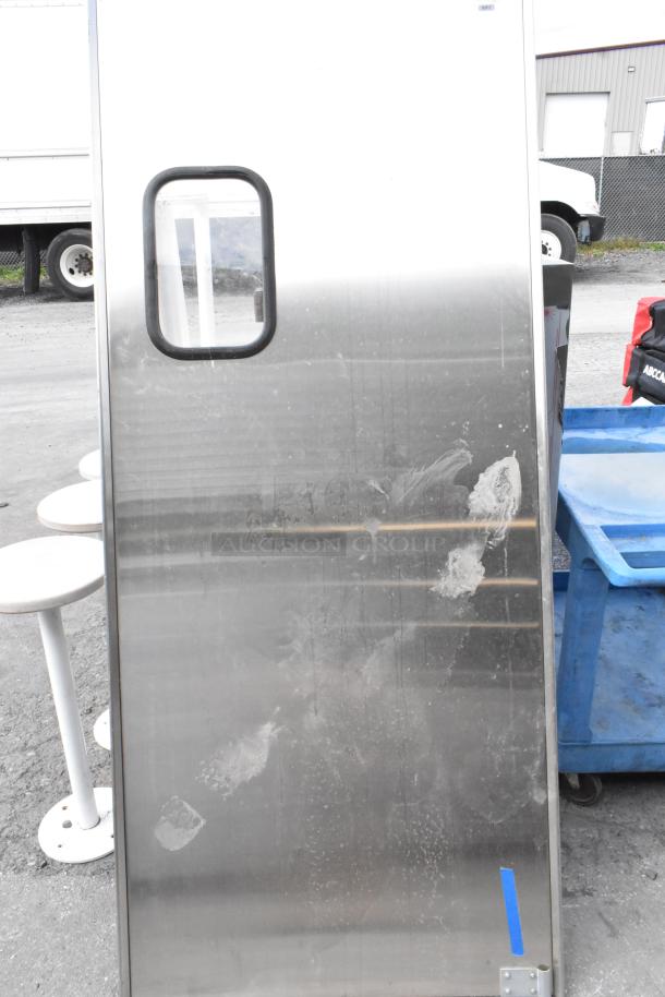 Stainless steel swinging kitchen door with rectangular window, showing wear. Durable for commercial kitchens.