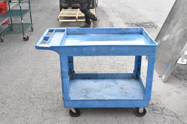 Blue two-tier poly cart on commercial casters, used condition, features handle and sturdy shelves, ideal for transport.