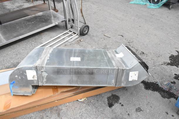 Metal duct with a rectangular shape, visible seams, and attached labels, indicating industrial-use application. Signs of wear visible.