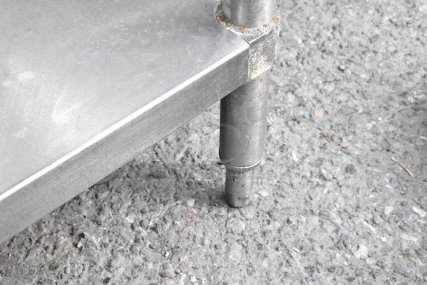 Stainless steel table corner with rusting under shelf; visible cylindrical legs; minor wear on surface.