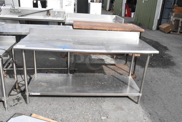Stainless steel table with back splash and under shelf. Features visible wear, sturdy frame, ideal for kitchens or workshops.