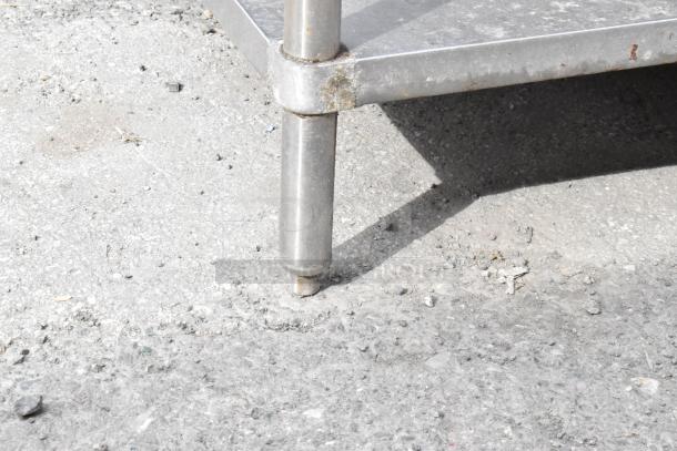 Stainless steel table corner with visible under shelf, showing moderate wear on leg joint, placed on rough concrete.