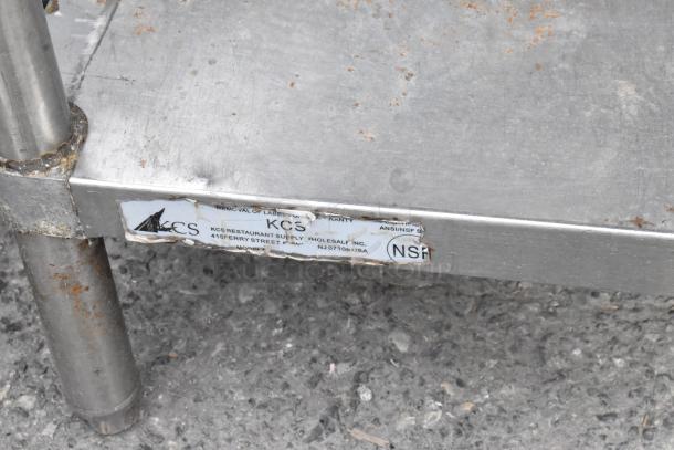 Stainless steel table with back splash and under shelf, visible wear, label from KCS Restaurant Supply showing NSF certification.