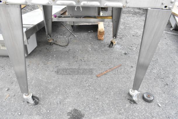 Hardt Inferno 4500 stainless steel rotisserie oven with skewers, metal legs, and commercial casters, shown outdoors.