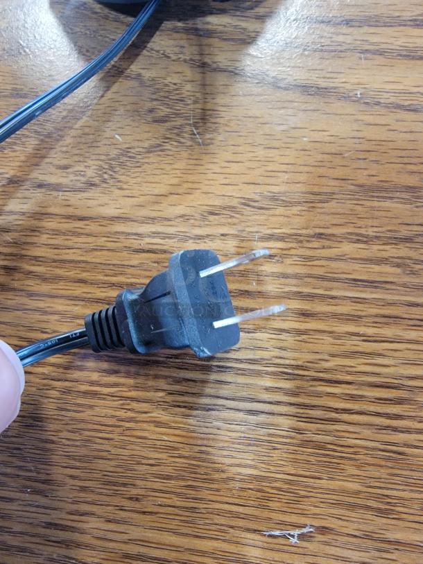 Close-up of a black AC power plug for the Element Lux Neon LED OPEN Sign, showing prongs and cable on wooden surface.