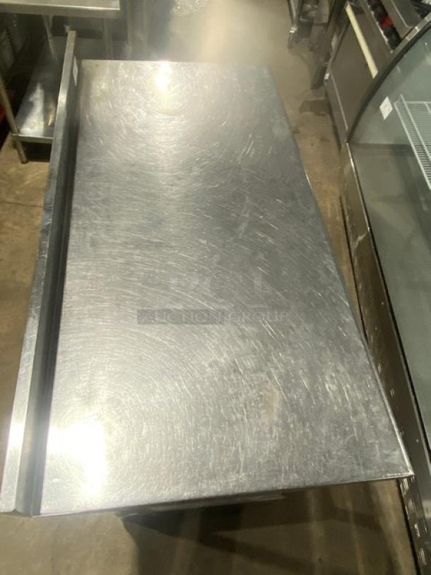 Commercial stainless steel worktop table with undershelf, up-turned backsplash, and legs. Visible wear, sturdy condition.