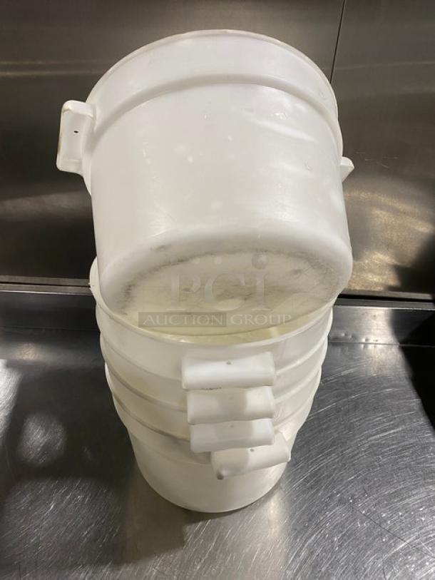 Commercial white round food storage containers with handles, stacked, showing signs of use.