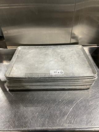 Stack of commercial baking sheet pans, worn condition, 12 pans total, labeled "80 X 12," stainless steel surface.
