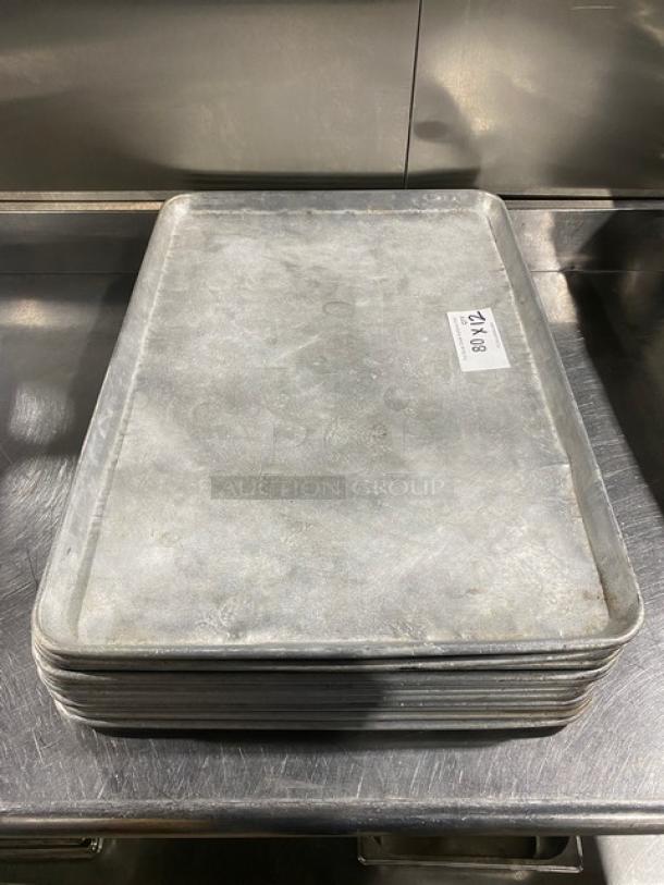 Stack of commercial baking sheet pans, silver, used condition, auction lot tag visible, 12 pans total, approx. 18x26 inches.