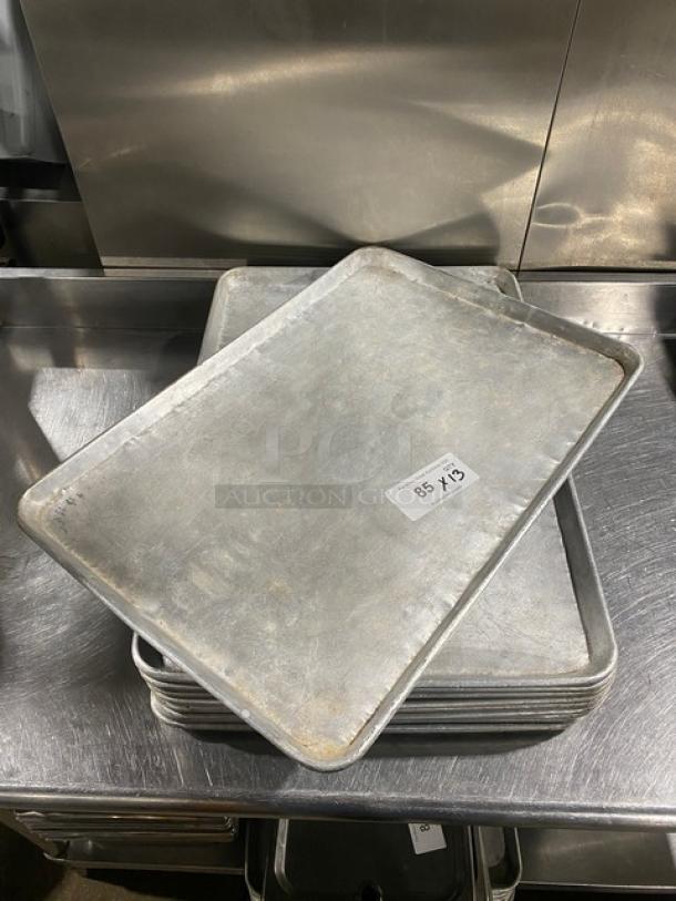Commercial baking sheet pans stacked on a stainless steel surface, showing moderate wear; labeled "85 X13."