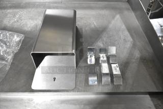 Stainless steel bin with a matte finish, featuring a keyhole on the front. Includes four metal clips with brand tags visible.