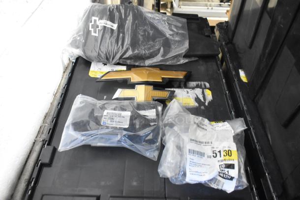 Lot of brand new car parts, including Chevy emblems, in a black poly bin. Items in sealed packaging with visible labels.