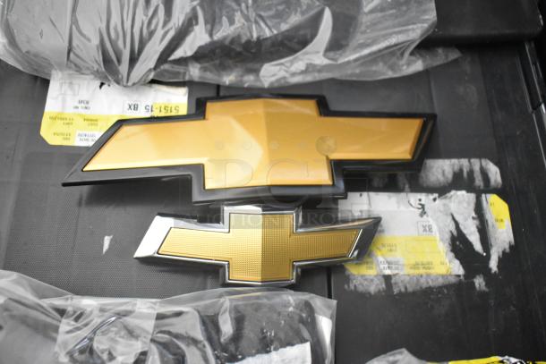 Brand new lot of car parts including two gold Chevy emblems in a black poly bin. Emblems pristine with protective packaging.