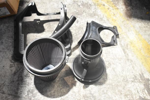 Lot of new car parts including air filter and black components, marked "ROUSH," on concrete floor.