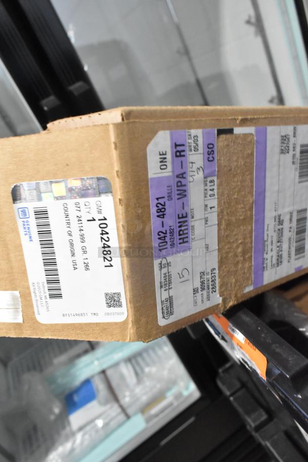 Brand new GM car parts in box, including GM 10424821 grille assembly, barcode and labels visible, in black poly bin.