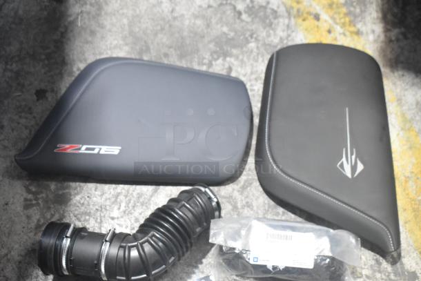 Brand new car parts including two black panels with logos, flexible black hose, and a bag with a barcode in a black poly bin.