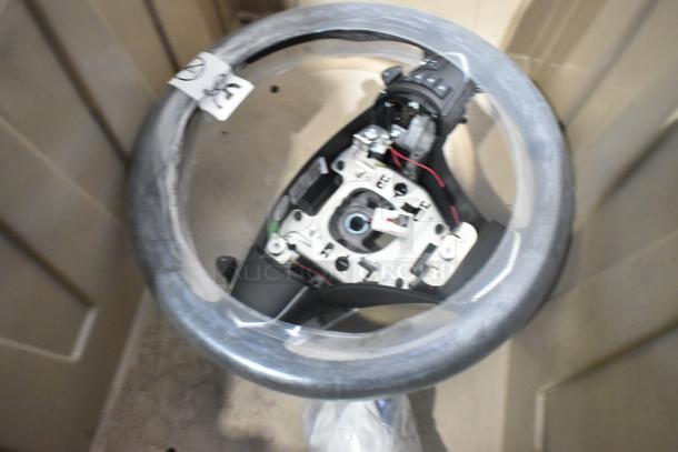 Brand new car parts lot in tan poly bin featuring a steering wheel with controls and tags attached.