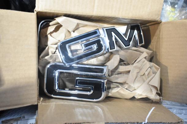 Brand new GM emblems in packaging, stored in a black poly bin with assorted car parts.