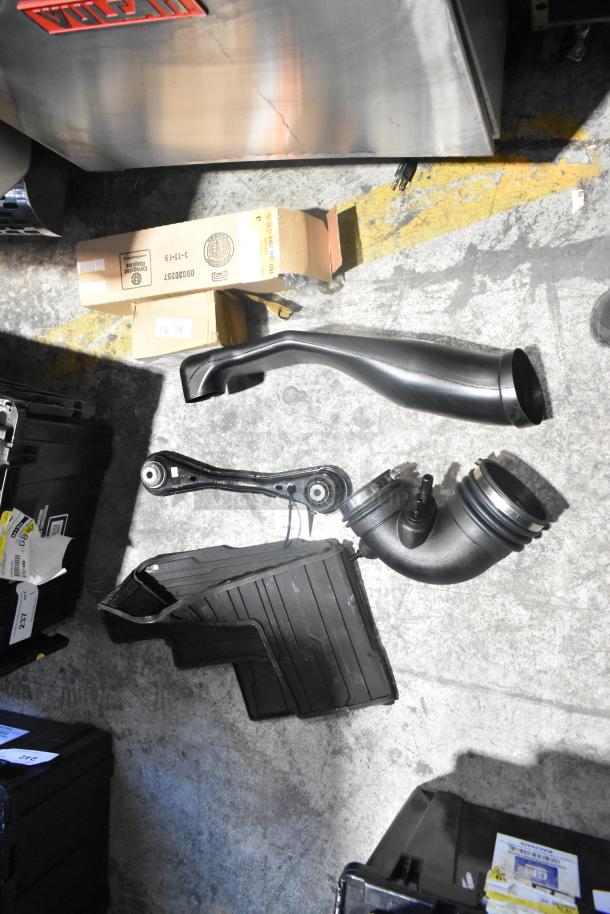 Brand new car parts lot including black car mats, air intake duct, and suspension arm, in a black poly bin. Boxes labeled.