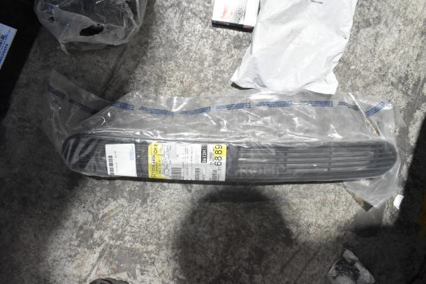 Brand new car grille in clear packaging on floor, labeled with part details, surrounded by other car item packaging.