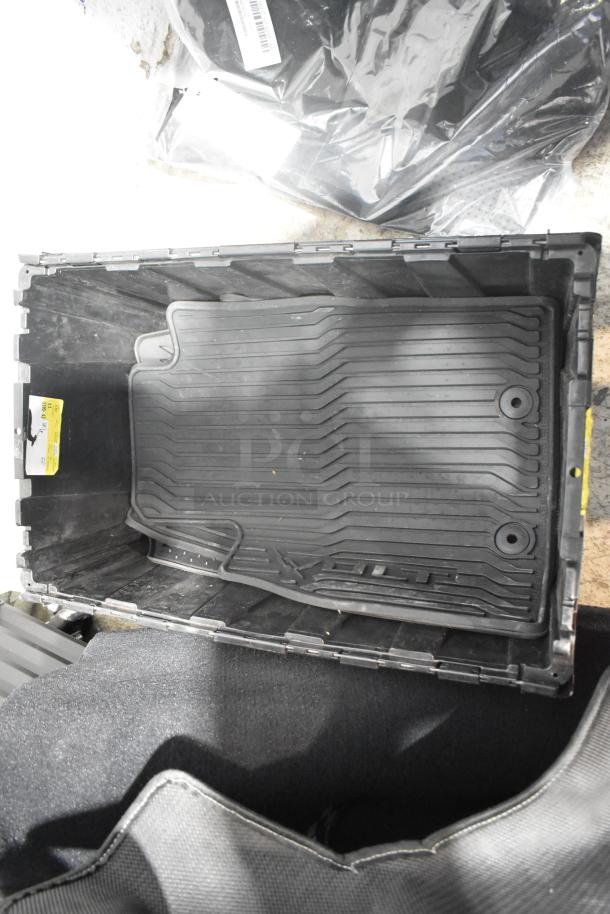 Black poly bin containing brand new car parts, including rubber car mats and an emblem, with visible labels and packaging.