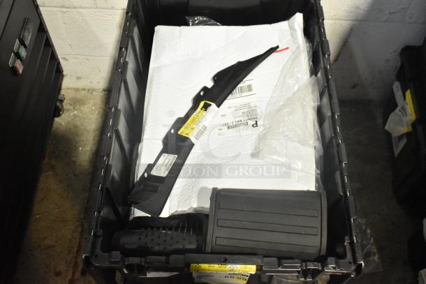 New car parts lot with mats, emblem, and items in black poly bin. Tags with barcodes and labels visible, indicating brand and specs.