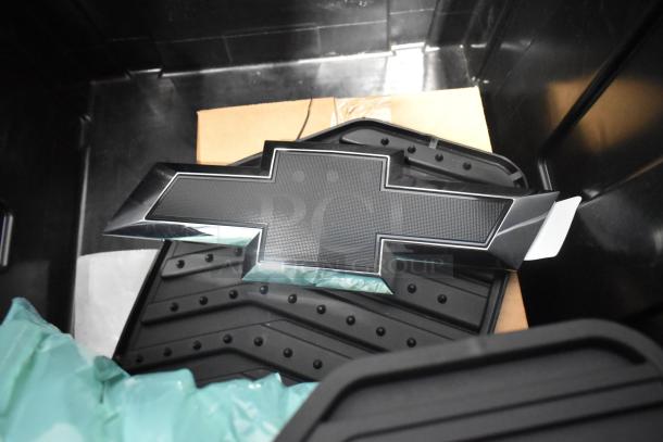 Brand new lot of car parts in black poly bin, includes car mats and a Chevrolet emblem.