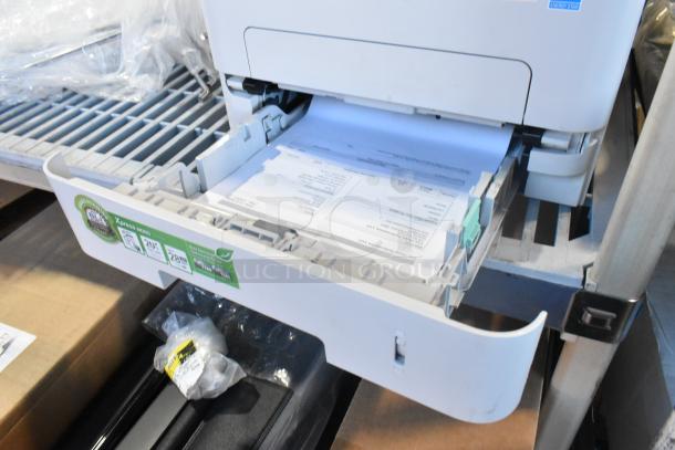 Samsung Xpress M2885FW printer with open paper tray, showing documents being printed. Condition appears functional.