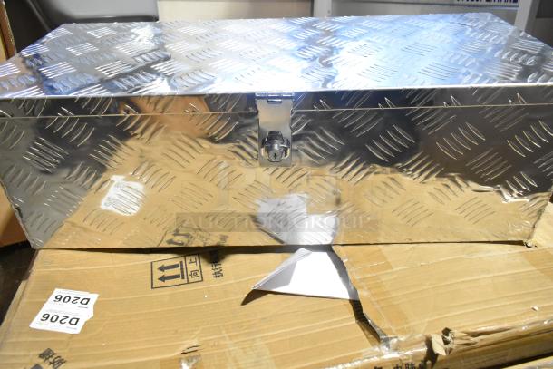Diamond plate tool box on cardboard, featuring a latch lock mechanism. Mild wear visible; box accompanies other storage items.