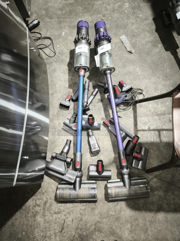 Dyson V11 Torque Drive PLUS cordless vacuum cleaners with multiple attachments, labeled 103 and 104, displayed on the floor.