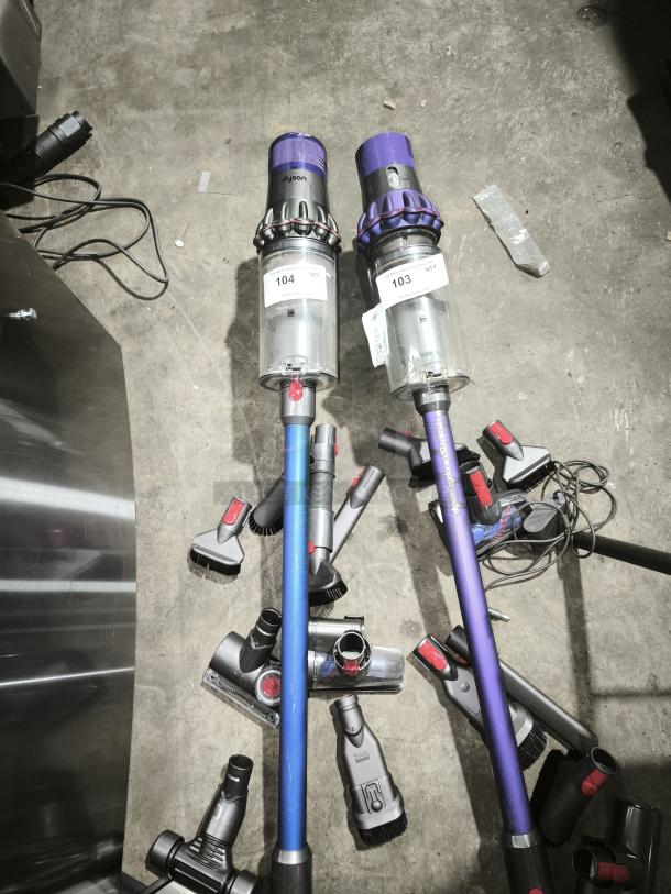 Dyson V11 Torque Drive PLUS cordless vacuum cleaners on floor, in used condition with attachments. Tags indicate auction lot numbers 103 and 104.