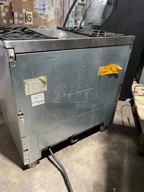 Monogram commercial stainless steel 4-burner gas range/oven combo. Features wired oven rack, set on sturdy legs. Visible caution labels.